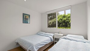 A bedroom with two beds, a small bedside table, a window with a view of greenery, and a framed picture on the wall. - Quat Quatta - Point Lonsdale (Point Lonsdale)