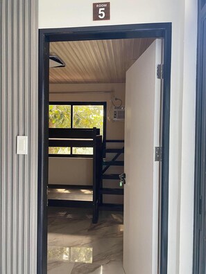 A room with a wooden bunk bed, a window with a view of greenery, and a sign indicating 'ROOM 5'.