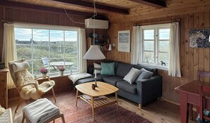 Een gezellig chaletinterieur met een bank, een klein tafeltje, een stoel en een raam met uitzicht op een natuurlandschap.