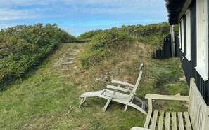 A wooden bench and a chair outside a building with a grassy area and a path leading uphill. - Family summer house on the dune with sea views (Ringkøbing)