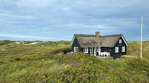 Une maison noire avec un toit de chaume, des fenêtres blanches et une cheminée, entourée de collines verdoyantes et sous un ciel dégagé.