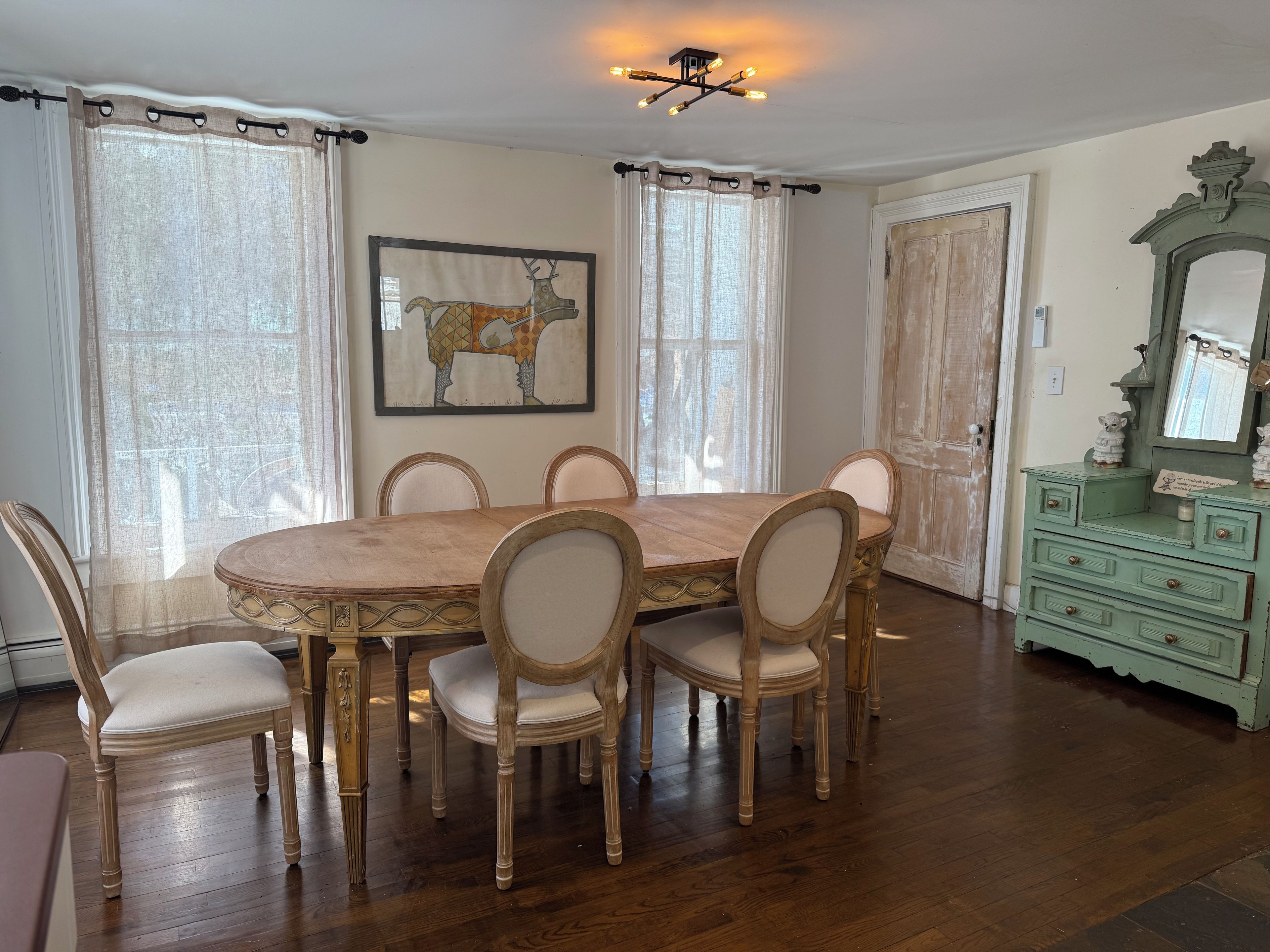 A dining area with a wooden table and chairs, a painting on the wall, and a dresser with a mirror.