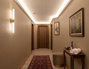 A hallway with a wooden door at the end, a patterned rug on the floor, a small round ottoman, and a table with towels and a vase. - Buyuk Paris Palace Hotel (İstanbul)