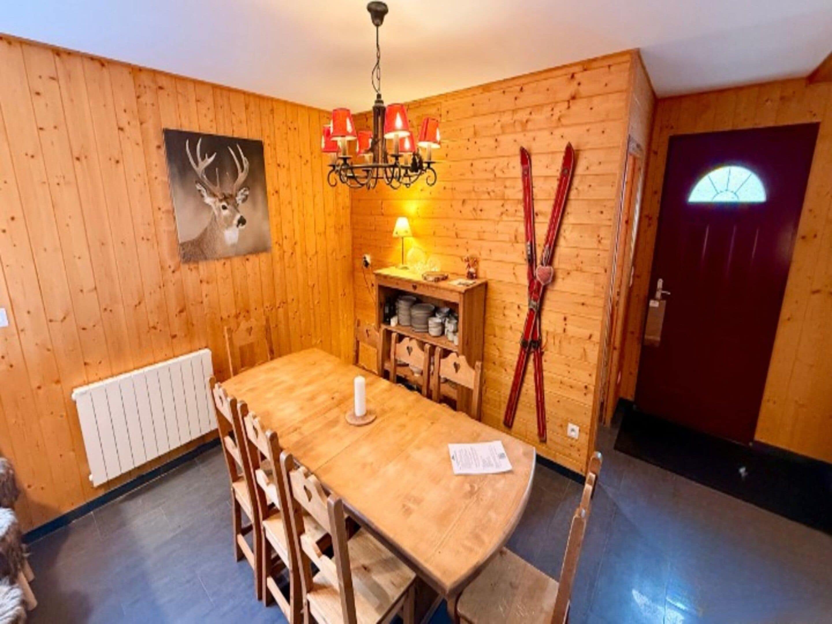 A wooden-paneled room with a dining table, chairs, a chandelier, a coat rack with skis, a door with a window, and a radiator.