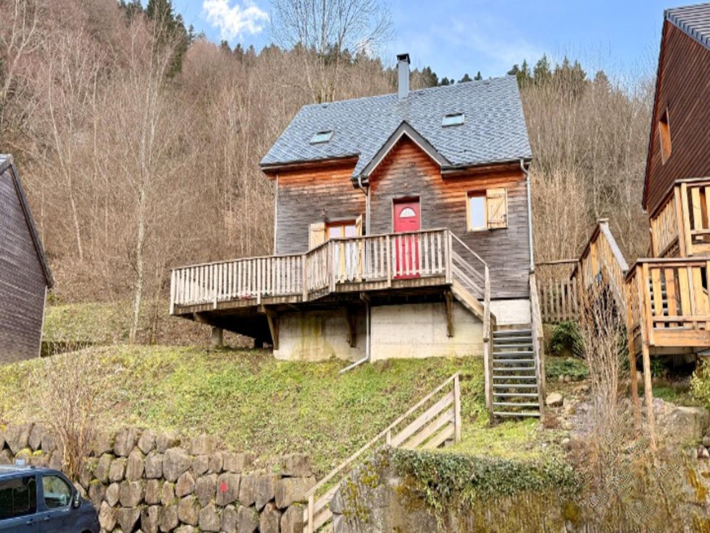 A wooden house with a balcony, a red door, and a stone wall in the foreground.