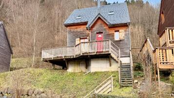 A wooden house with a balcony, a red door, and a stone wall in the foreground.