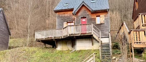 A wooden house with a balcony, a red door, and a stone wall in the foreground.