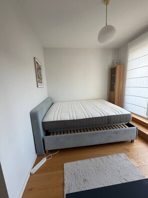 Een compacte kamer met een bed, een stekkerdoos, een klein tapijt en een raam met jaloezieën.