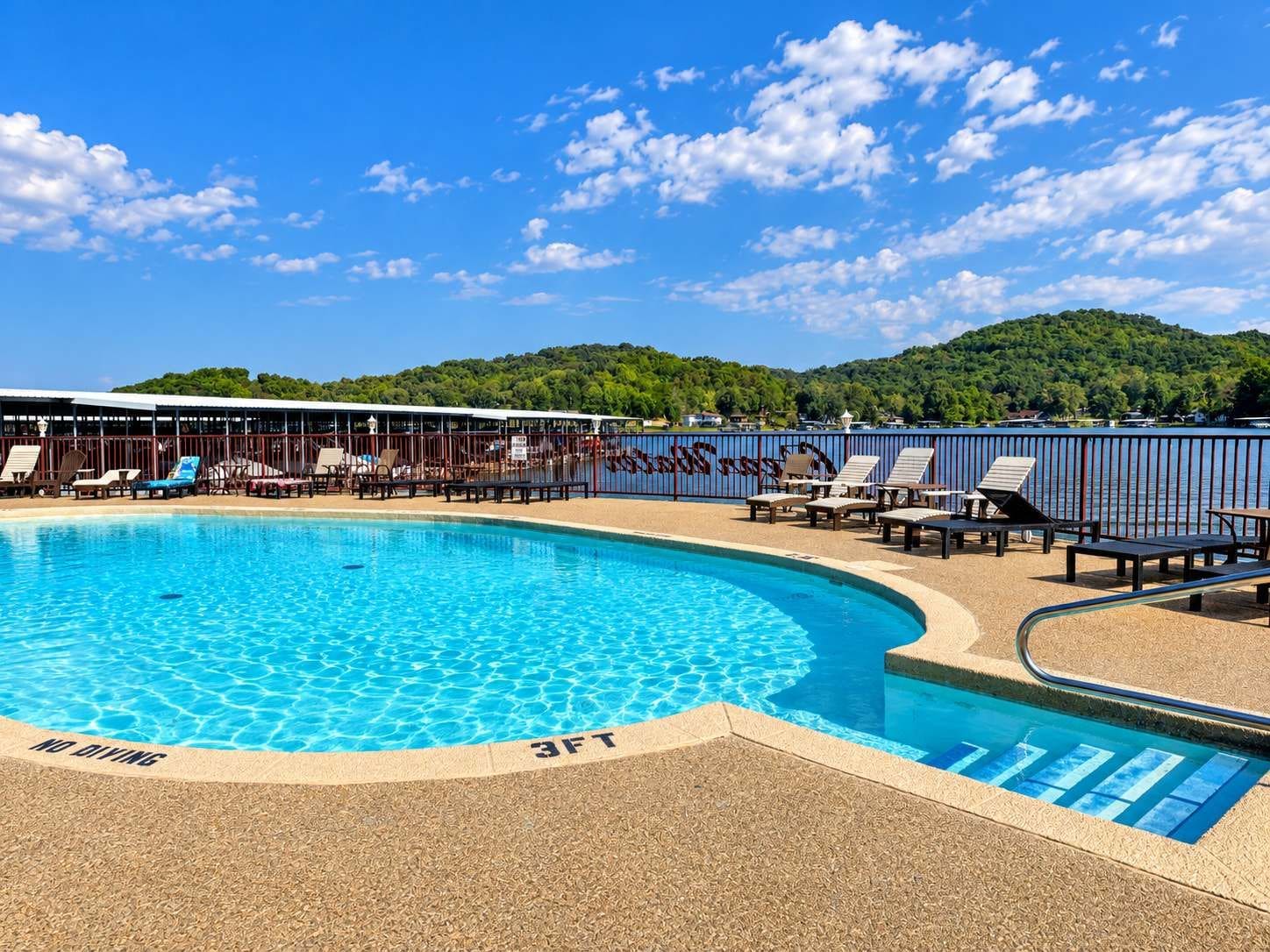 A swimming pool with lounge chairs and a no diving sign, surrounded by a fence and a view of a forested hill.