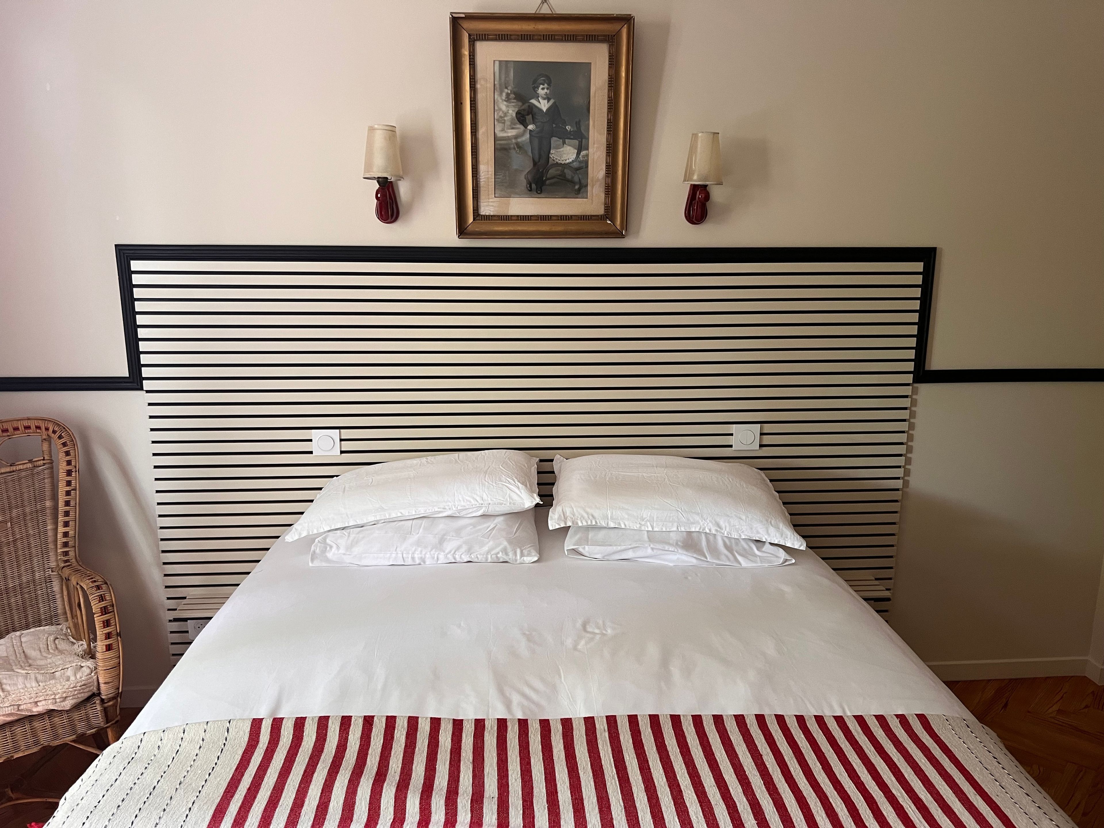 Ein Bett mit weißen Leinentüchern und einer gestreiften roten und weißen Decke, ein Kopfteil mit horizontalen Linien sowie zwei an der Wand befestigte Lampen.