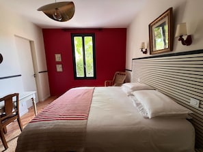 Ein Schlafzimmer mit einem Bett, einem Sessel, einem Schreibtisch, einem Fenster und einer Wandlampe.