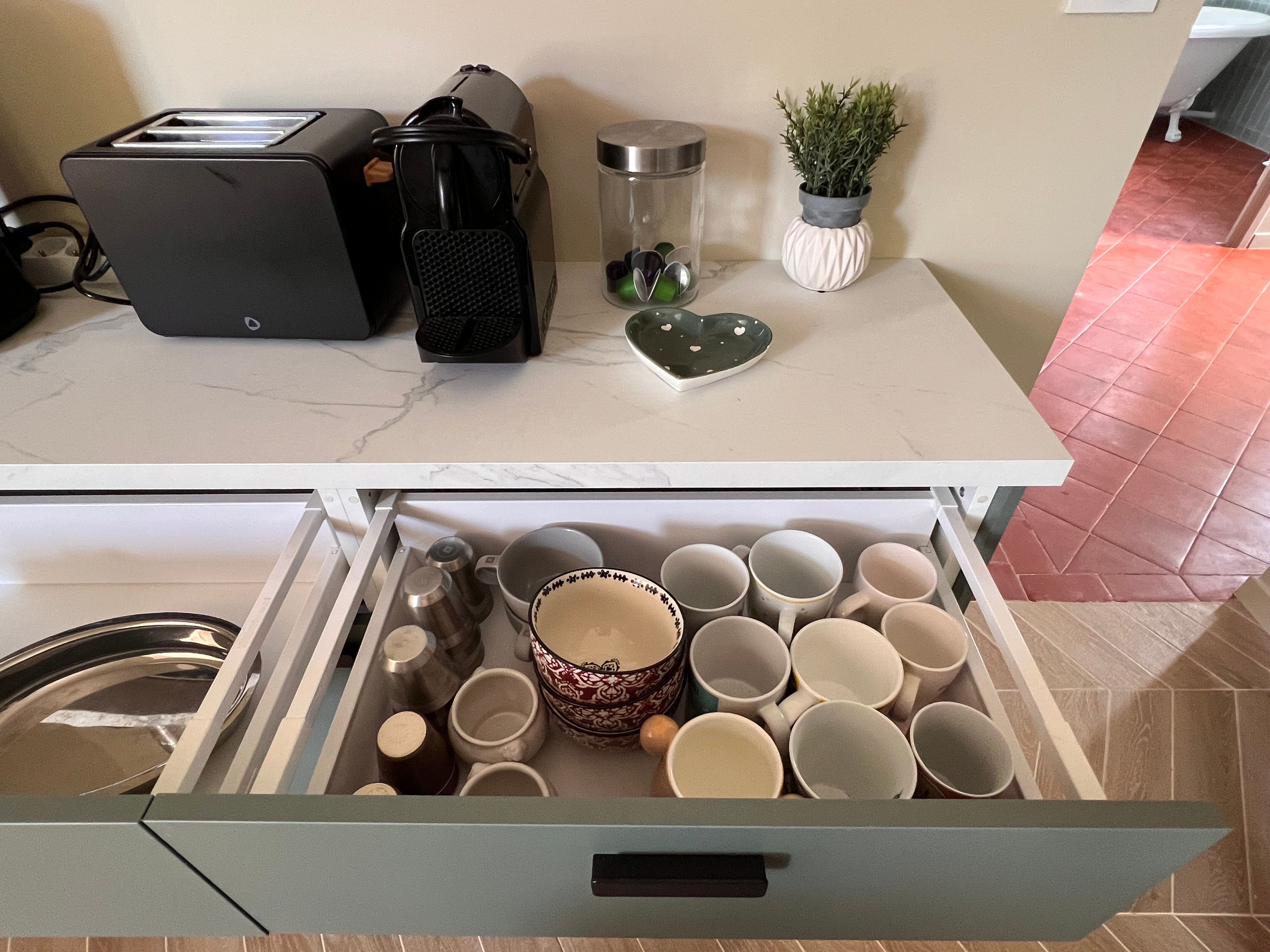 Eine Küchenarbeitsplatte mit einer Kaffeemaschine, einer Pflanze und einem herzförmigen Gegenstand. Darunter befindet sich eine Schublade mit verschiedenen Tassen und Behältern.