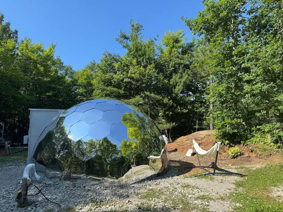 A spherical structure with a reflective surface, surrounded by trees and a gravel area.