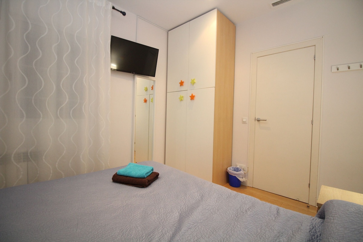 A bedroom with a bed, a wardrobe, a television, and a trash bin.