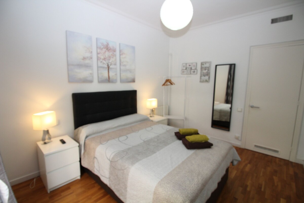 A bedroom with a bed, bedside tables, a nightstand, a mirror, and wall art.