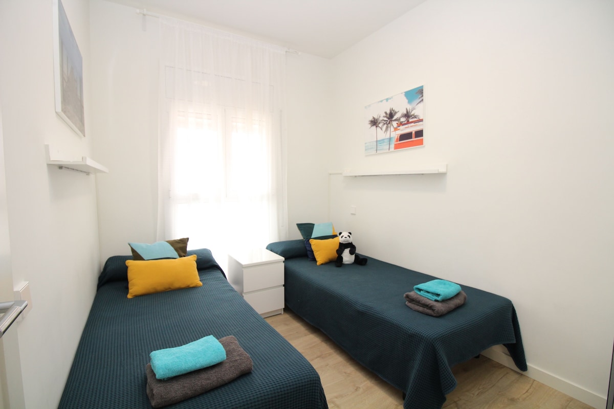 A room with two single beds, each with a pillow and a blanket. There is a window with sheer curtains, a wall-mounted picture, and a small shelf with a picture.