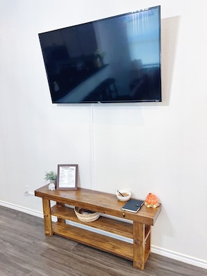 A flat-screen TV mounted on a wall above a wooden console table with a framed welcome sign, a small plant, and a decorative pumpkin. - The Blue Cottage- 3BR Family Retreat on Lake Granbury, Gated, Community Pool (Granbury)