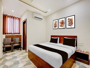 A hotel room with a bed, a nightstand, a chair, a wardrobe, and a window with curtains. - Deluxe Room Hotel Shivalika (Jaipur)