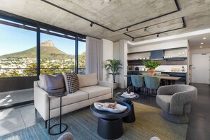 A modern living room with a sofa, coffee table, and chairs, a kitchen area with bar stools, and a large window with a view of a hill. - FortyOnL | Stunning City and Table Mountain Views (Cape Town)