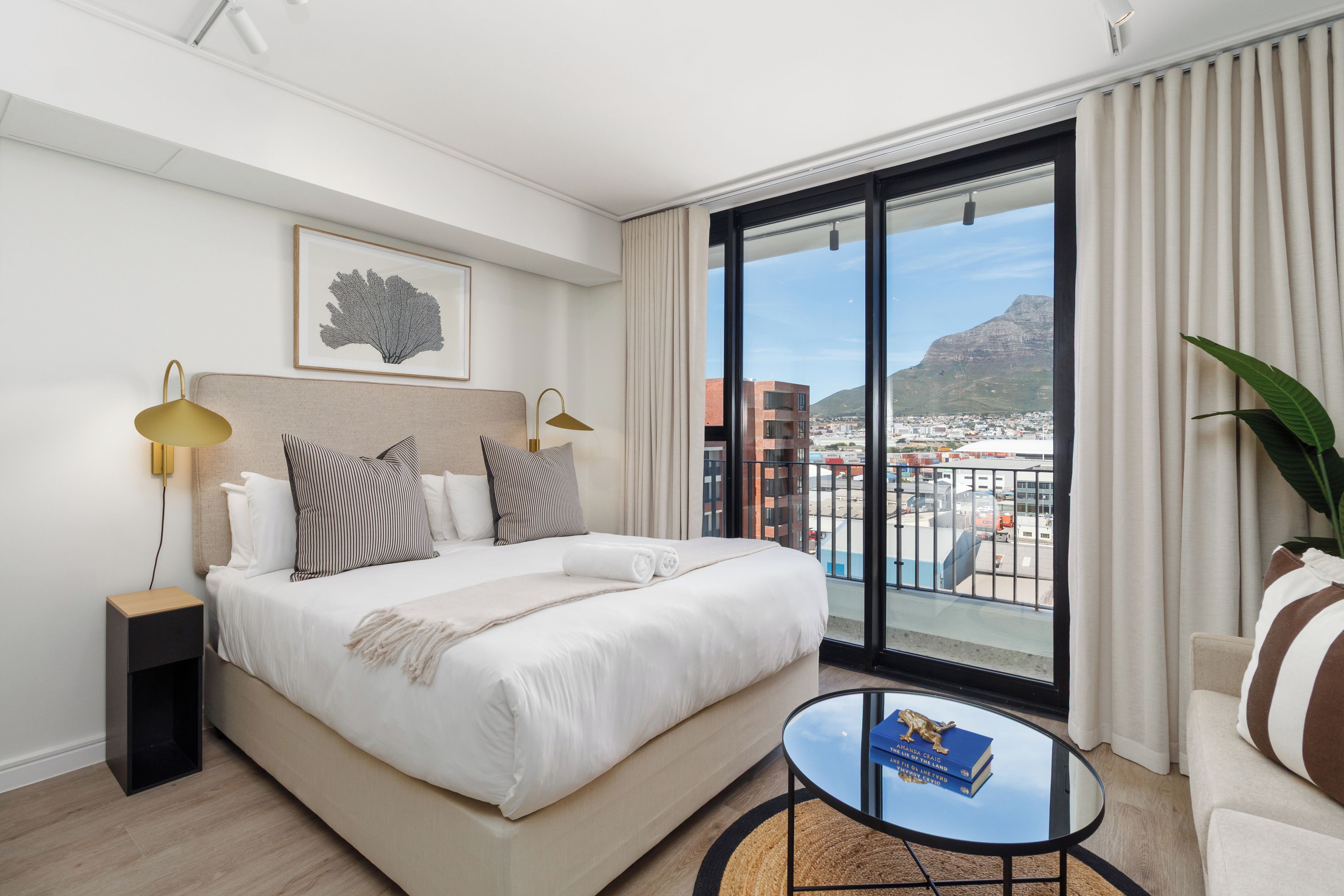 Habitación de hotel con cama, lámparas de noche, vistas a la ciudad y una pequeña mesa con libros.