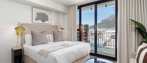 Habitación de hotel con cama, lámparas de noche, vistas a la ciudad y una pequeña mesa con libros.