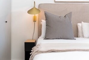 A neatly made bed with white bedding and striped pillows, a bedside lamp, and a small wooden nightstand. - Stylish City Escape | Rooftop Pool and Gym (Cape Town)