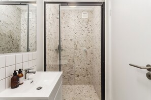 A bathroom with a white sink, a mirror, a shower with a glass door, and a tiled floor. - Stylish City Escape | Rooftop Pool and Gym (Cape Town)