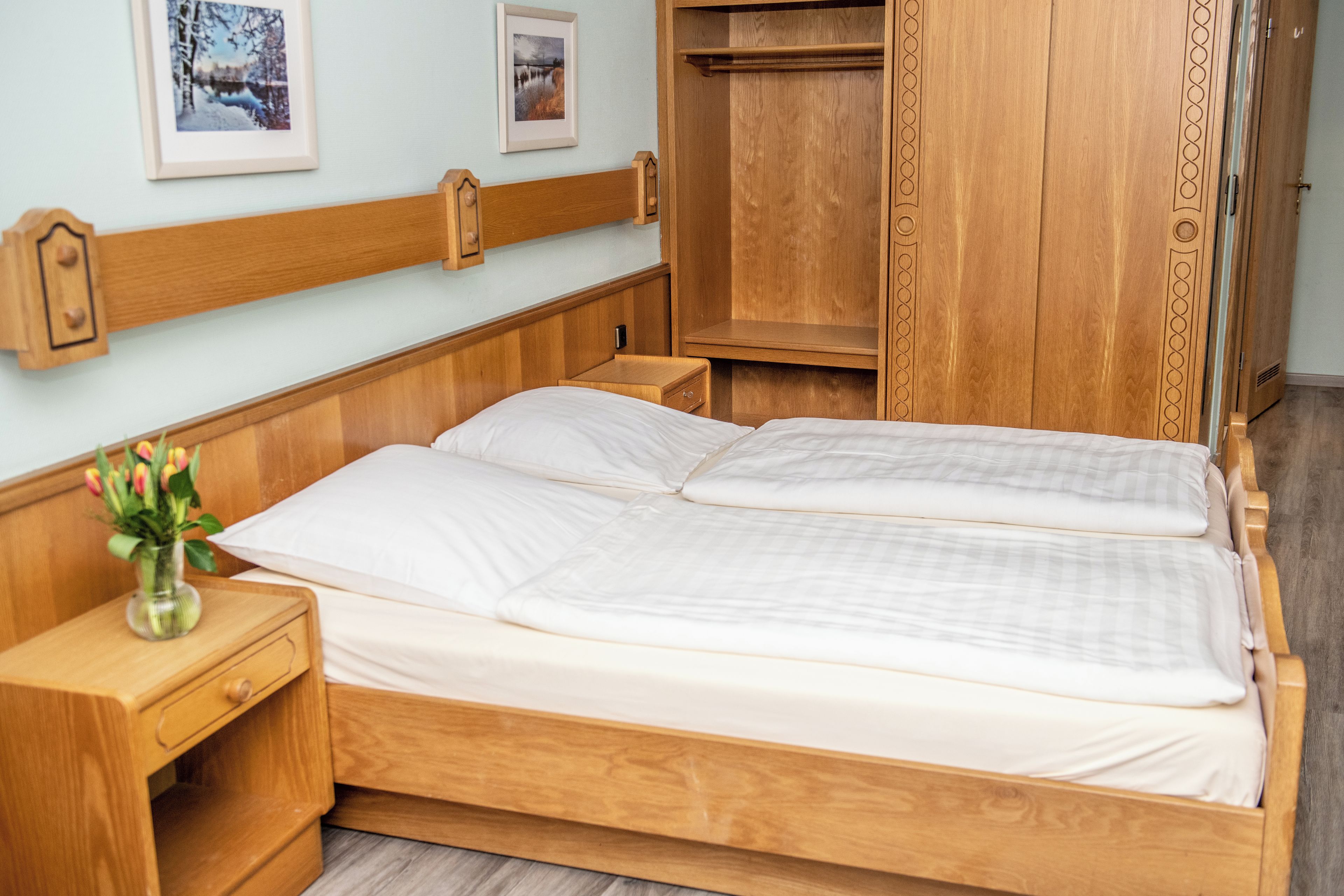 Ein hölzernes Bett mit zwei Kissen, ein Nachttisch mit Blumenvase und ein Kleiderschrank mit Spiegel.