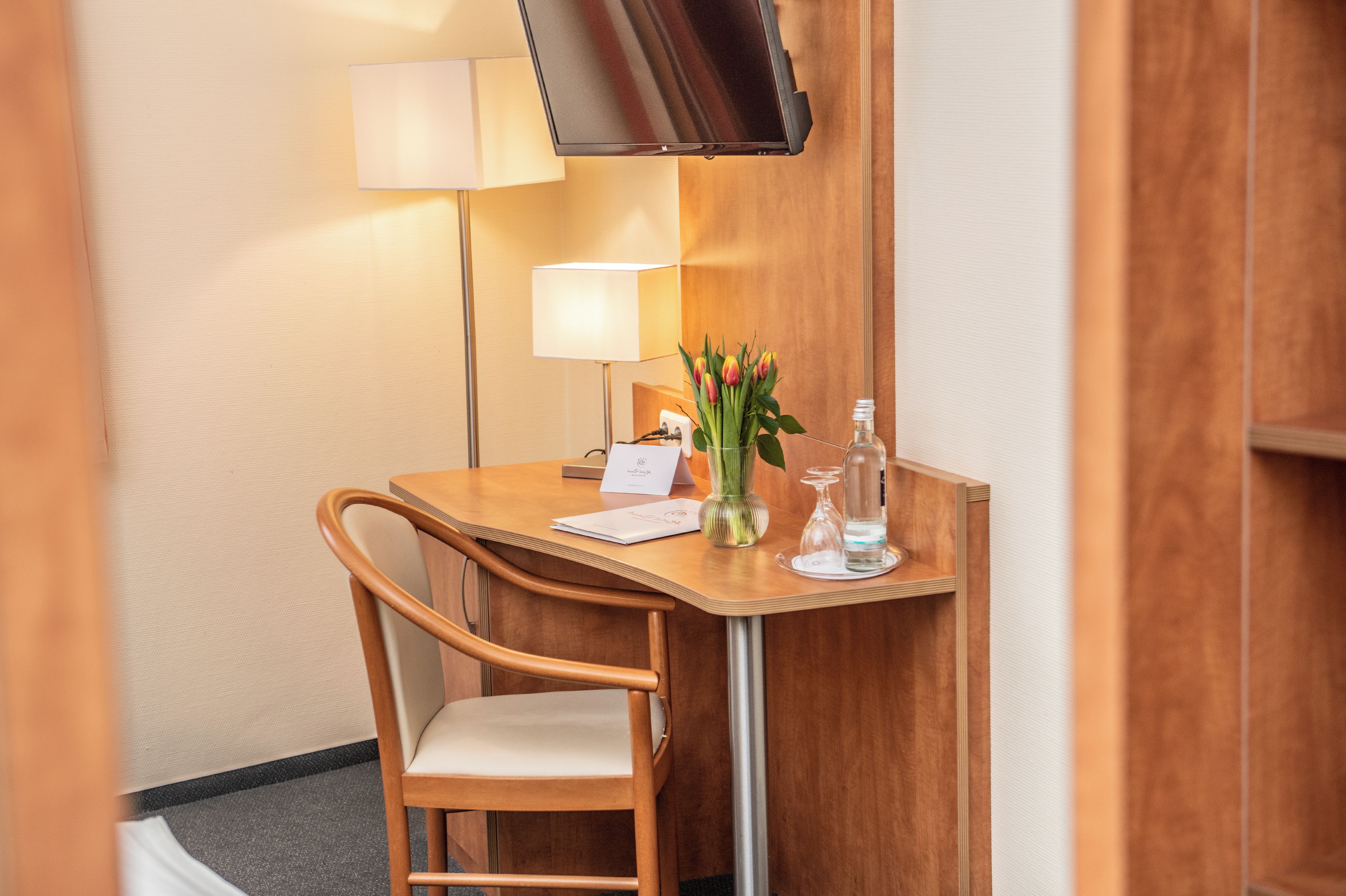 A hotel room with a wooden desk, a chair, a flat-screen TV mounted on the wall, a lamp, a vase with flowers, and a bottle of water.