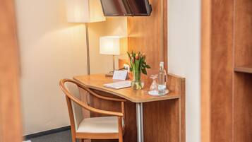 A hotel room with a wooden desk, a chair, a flat-screen TV mounted on the wall, a lamp, a vase with flowers, and a bottle of water.