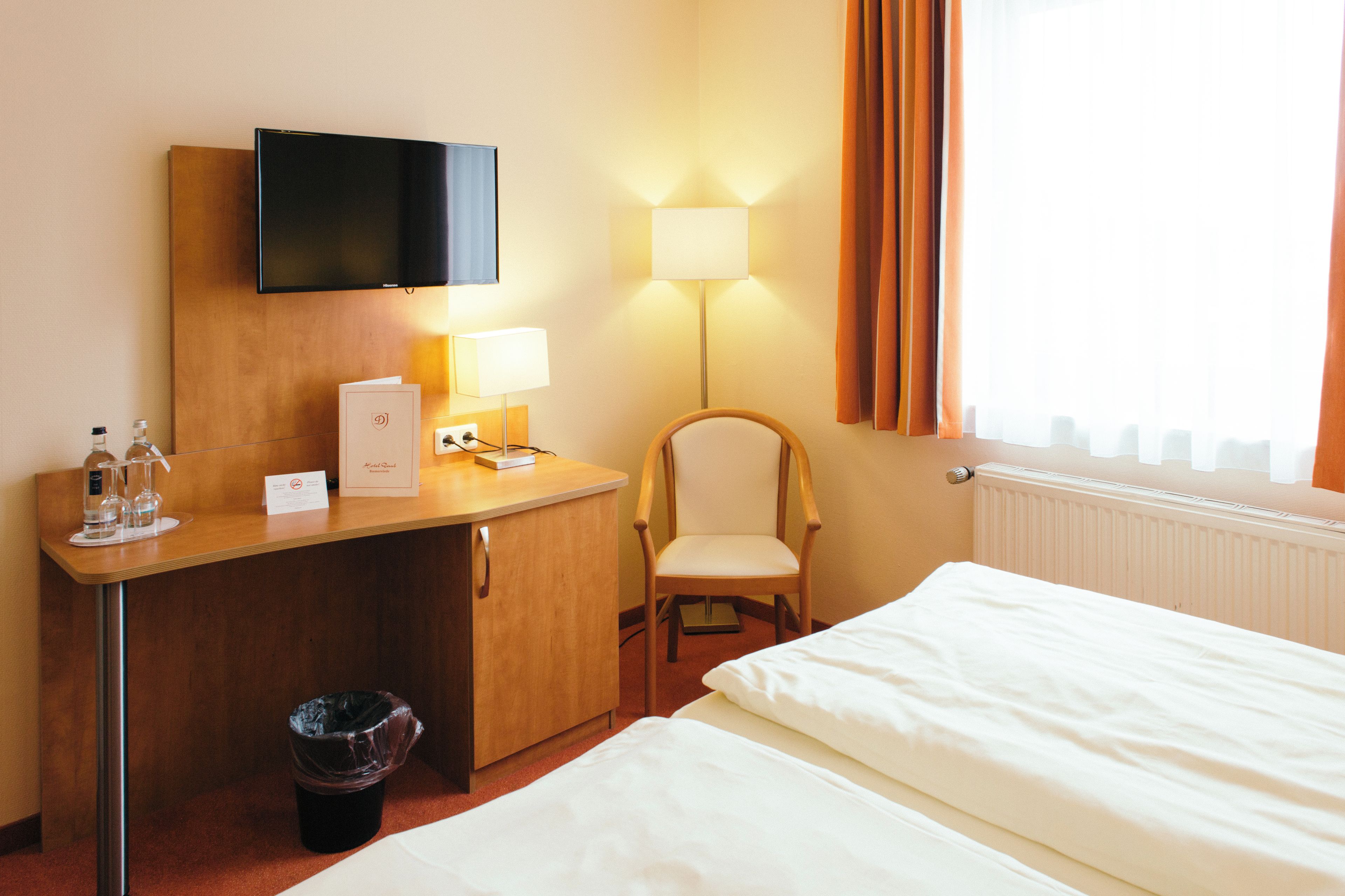 A hotel room with a bed, a desk with a television, a chair, a lamp, and a window with curtains.