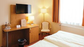 A hotel room with a bed, a desk with a television, a chair, a lamp, and a window with curtains.