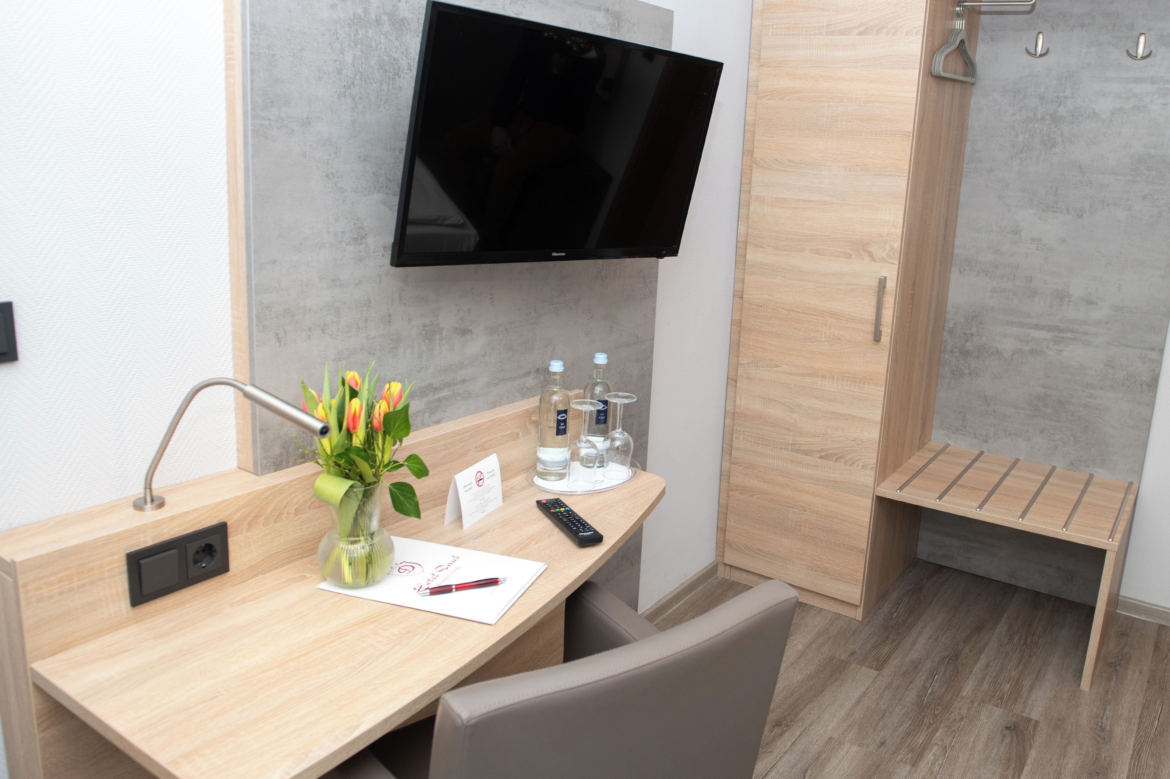 Una habitación de hotel con un escritorio de madera, un televisor en la pared, un florero con flores, una botella de agua y un vaso.