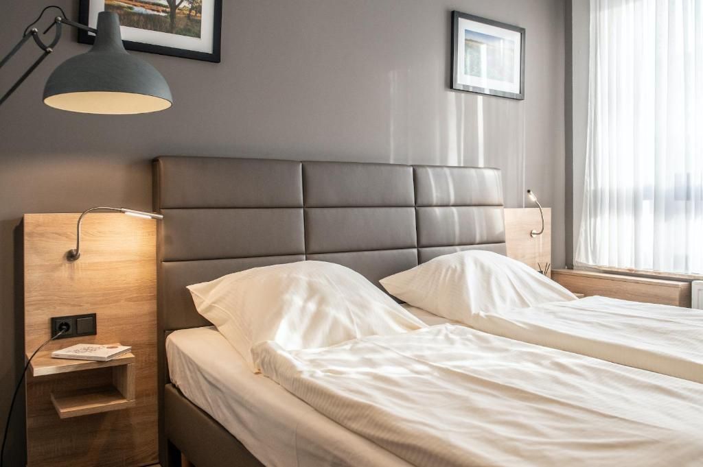 Habitación de hotel con dos camas, una mesita de noche, una lámpara y un ventanal con cortinas.