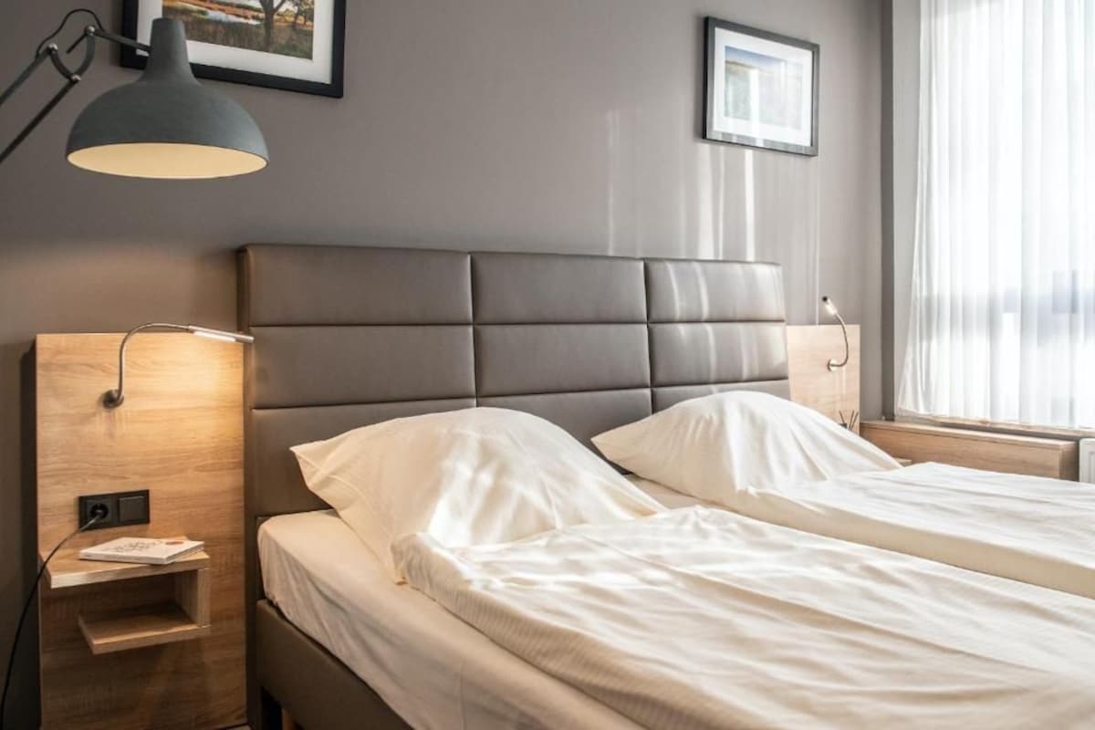 Quarto de hotel com duas camas, uma mesa de cabeceira, abajur e janela com cortinas.