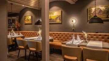 L’intérieur d’un restaurant présente un long banc en cuir marron, des tables rondes recouvertes de nappes blanches et un éclairage suspendu.