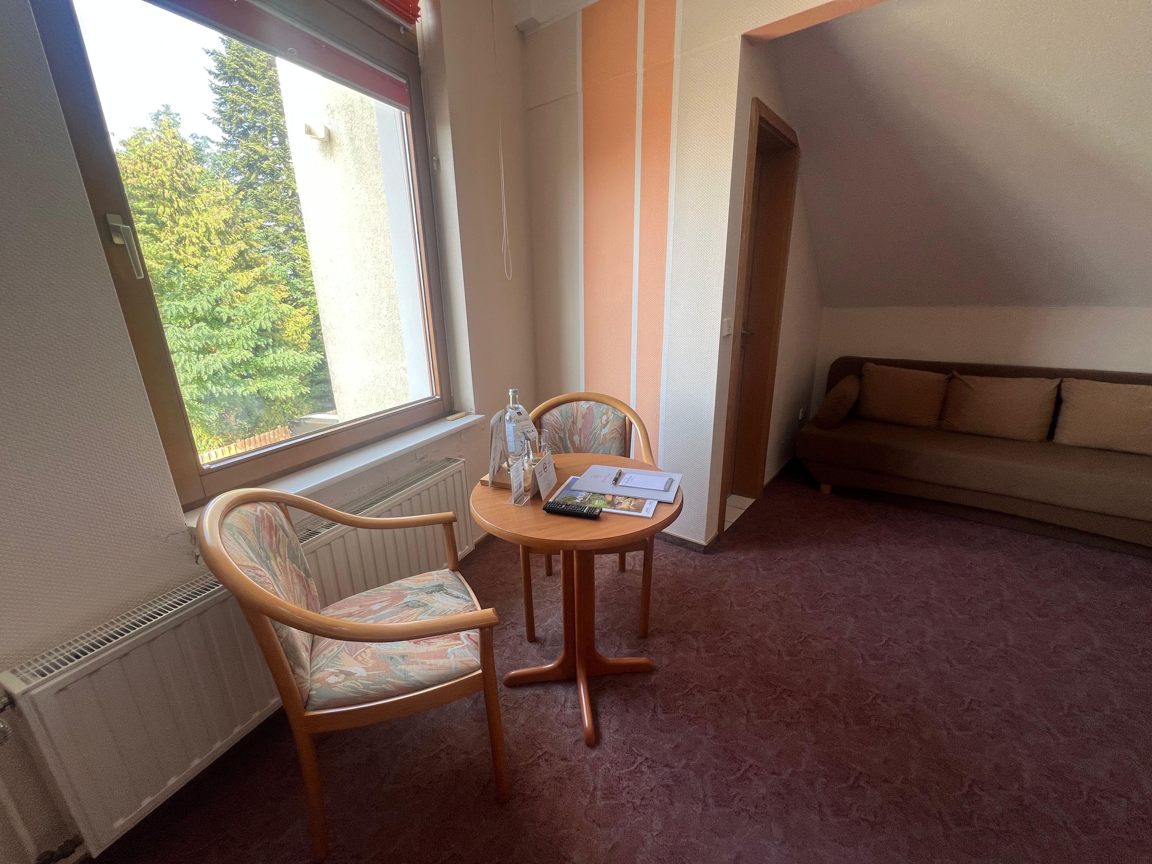 Una habitación con ventana, un radiador, un sofá, una mesa con sillas y suelo alfombrado.