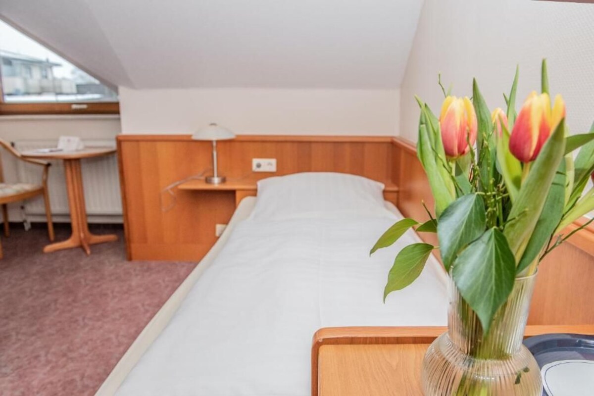 Une chambre d’hôtel avec un lit simple, une table de chevet en bois, une petite table avec une lampe, une chaise et un vase avec des fleurs.