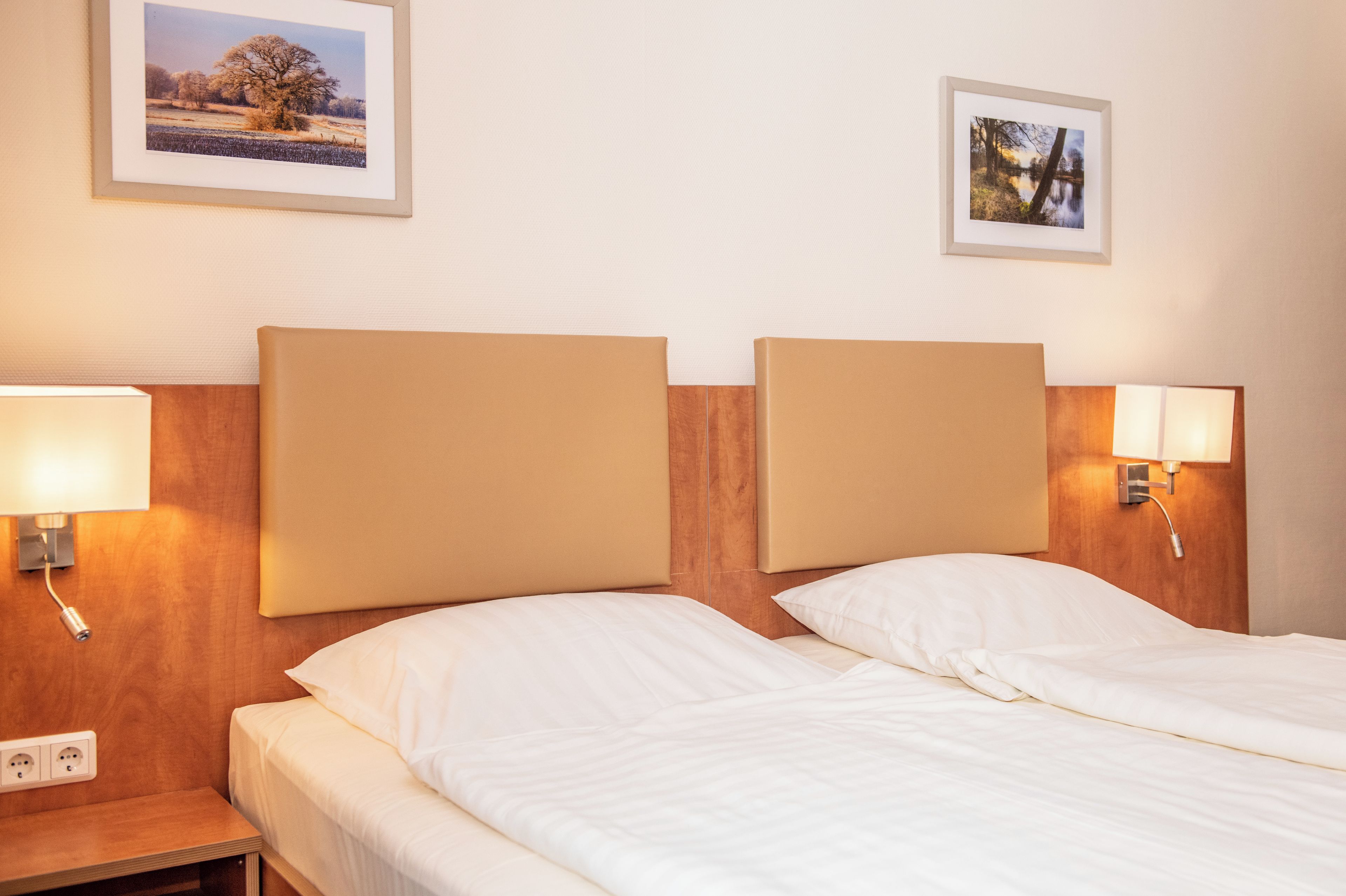 A hotel room with two beds, headboards, bedside lamps, and two framed pictures on the wall.