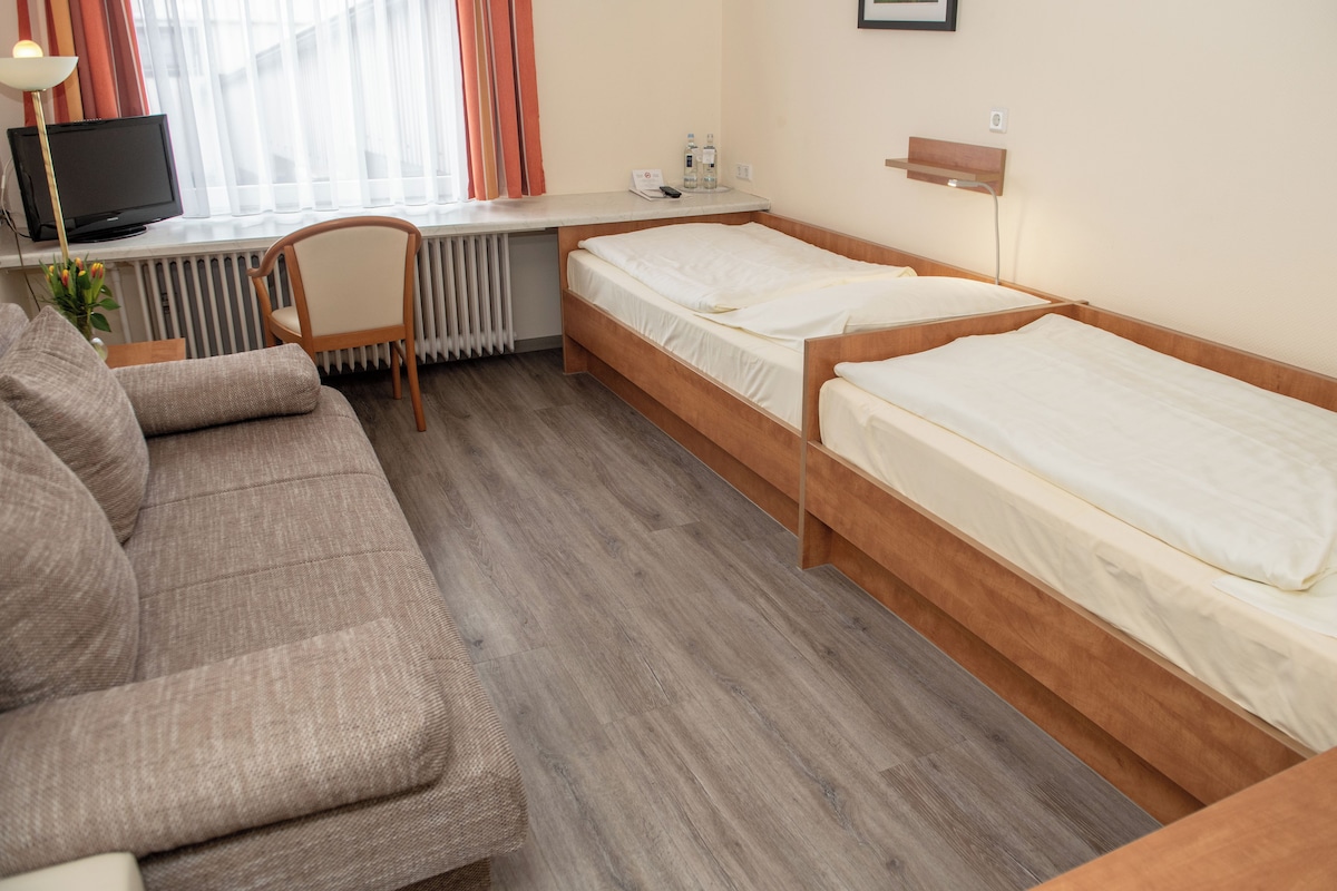 Quarto de hotel com sofá, duas camas de solteiro, escrivaninha com computador e radiador.
