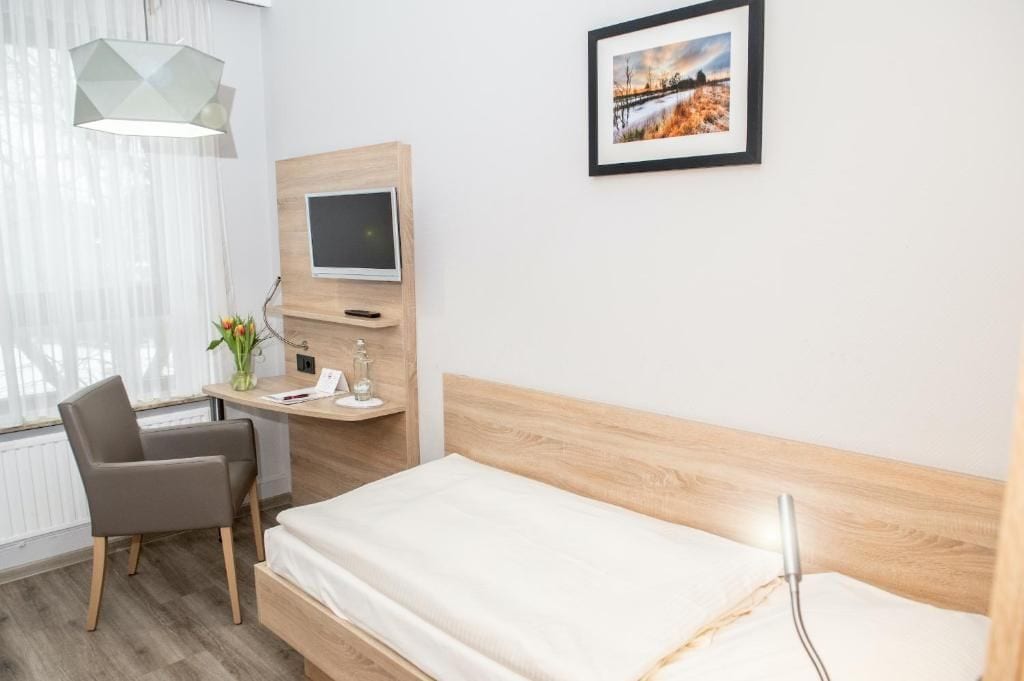 Una habitación de hotel con una cama, un escritorio con computadora, una silla, una ventana con cortinas y un cuadro en la pared.