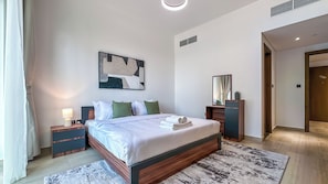 A modern hotel room with a large bed, bedside tables, a nightstand, a wall-mounted artwork, and a door leading to another room. - Modern 1BR apartment in Elysee 3 by Pantheon, Pool & Gym access (Dubai)