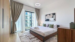 A modern hotel room with a large bed, a wooden dresser, a view of the cityscape, and a contemporary art piece on the wall. - Modern 1BR apartment in Elysee 3 by Pantheon, Pool & Gym access (Dubai)