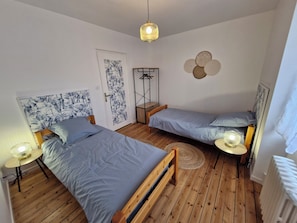 Un dormitorio con dos camas individuales, suelo de madera, una puerta con estampado floral, una lámpara colgante y un radiador fijado a la pared.
