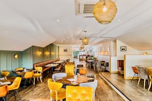 Een restaurantinterieur met houten tafels, gele en oranje stoelen en hangende lampen.