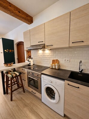 A modern kitchen with light wood cabinets, a stainless steel range hood, a built-in washing machine, and a black countertop. - Vrbo Property (Toulouse)
