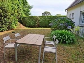 Une table en bois, entourée de quatre chaises, se trouve dans un jardin, au milieu de la verdure, avec un bâtiment blanc en arrière-plan.