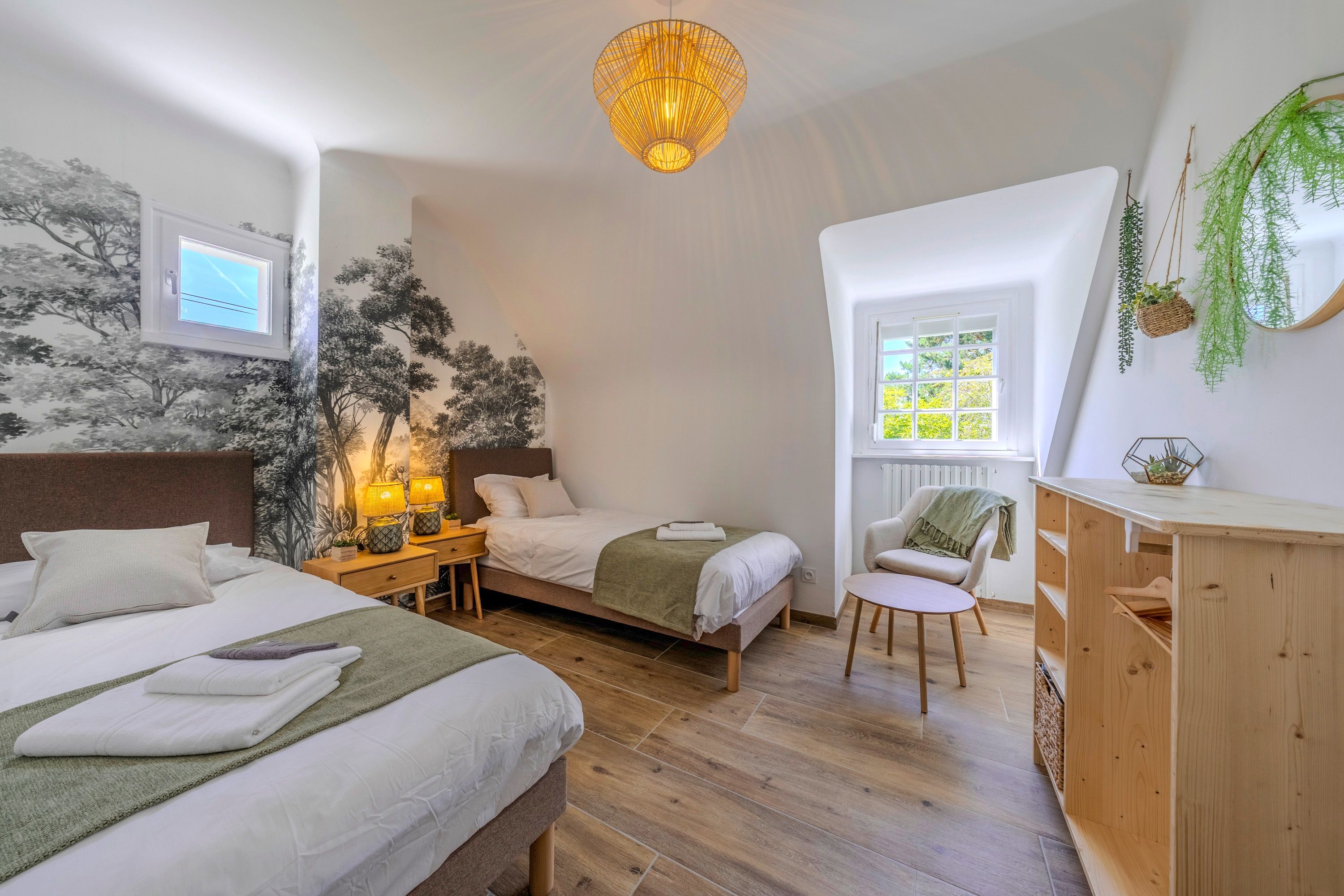 A bedroom with two beds, a desk, a chair, a hanging light fixture, and a window with a view.