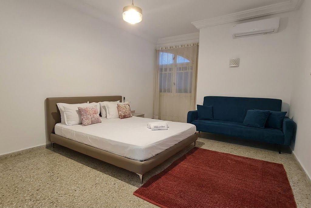 Un dormitorio con cama, un sofá azul, un tapete rojo y una ventana con cortinas.