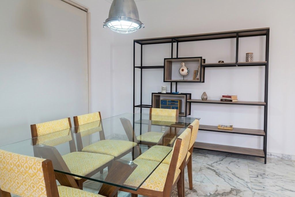 A dining area with a glass table and chairs, a metal-framed bookshelf, and a pendant light.
